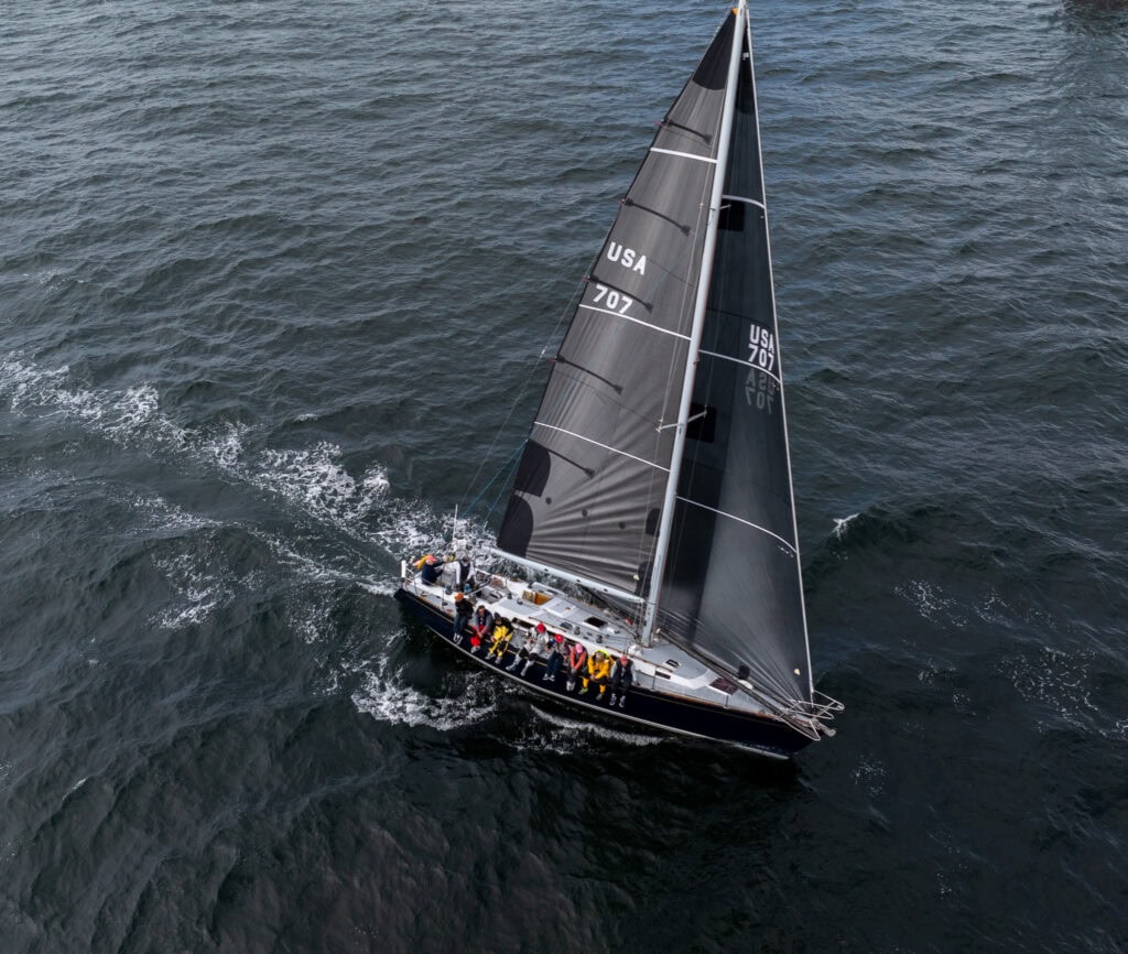 Aerial view of a dark racing sloop under sail with crew hiked out on the rail