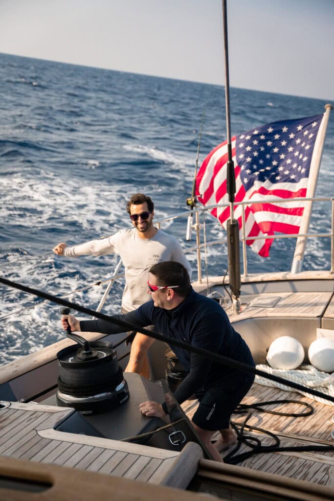 Two crew working a winch in the cockpit of a sailing yacht under way, United States ensign flying astern